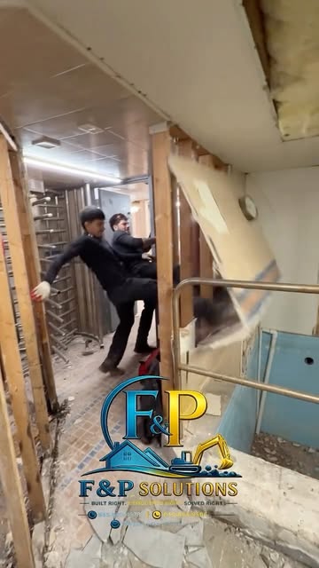 Two of our crew knocking out an interior wall during a demolition, broken wallboard and tile across the floor, the F&P Solutions logo in the corner.