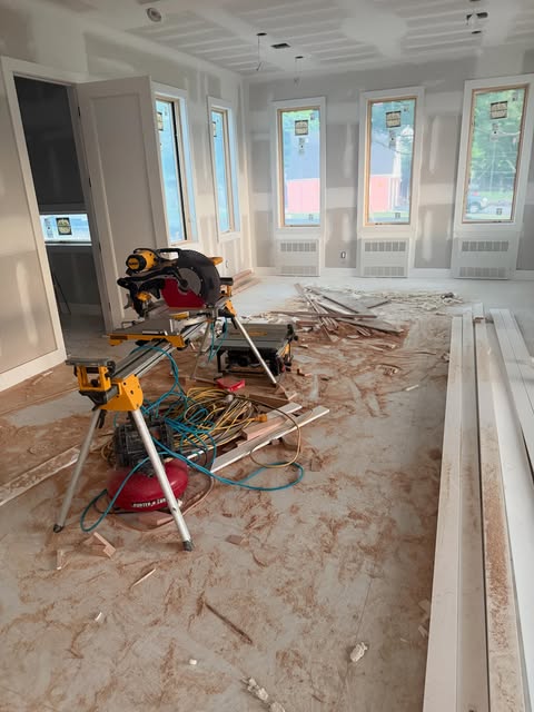 A bright new room with taped drywall and tall windows, a yellow miter saw on its stand and trim boards laid out on the floor.