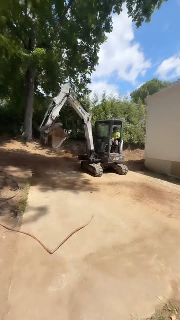 A mini excavator with an operator in a hi-vis shirt working a freshly graded lot beside a building, tall trees behind.