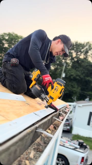 One of our crew in red gloves nailing down the edge of a roof with a coil nailer, trees and a pickup below.
