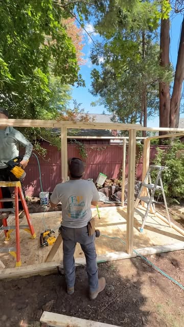 Two of our crew framing a custom shed on a new wood deck in a backyard, one in an F&P Solutions shirt, a red fence and trees behind.