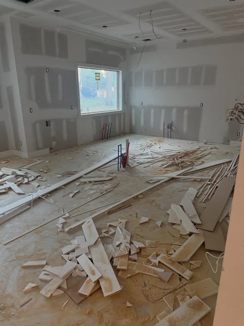 An unfinished room with taped drywall and a large window, cut trim pieces across the subfloor.